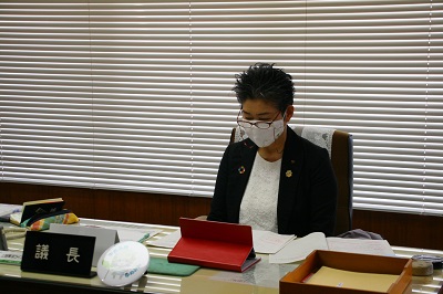 縦長の机が置かれ、灰色のスーツを着た短髪の女性が着座している。机上には赤いタブレット端末が置かれている