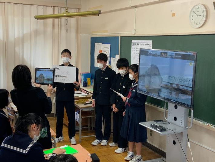 学校の教室で、黒い制服を着た男子生徒3人と女子生徒4人がいる。生徒の横にはテレビが置かれ、画面に議員の顔が映っている