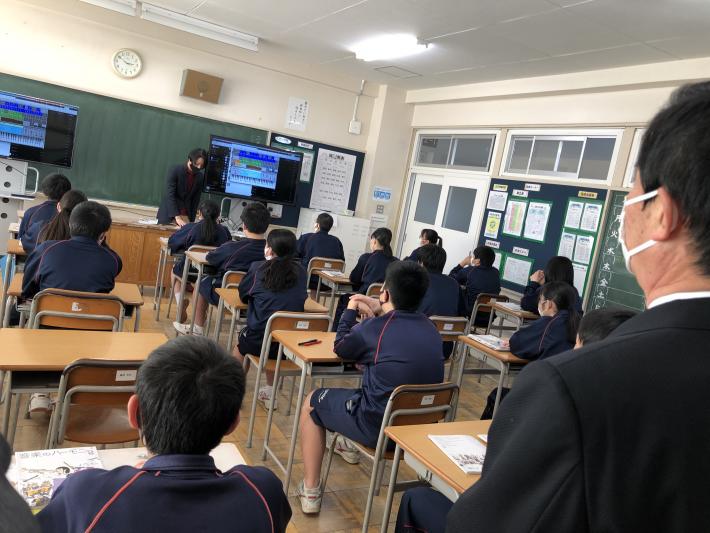 教室で生徒15人がこちらに背を向けて着席しながらパソコンの授業を受けている。一番手前にスーツの男性がこちらに背を向けて立っている