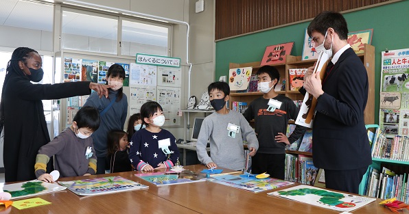 6人の子供が3人の先生と立ちながら机に置いてある絵本の表紙を見ている画像