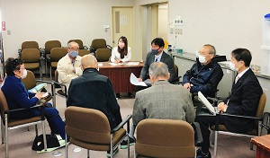 長い髪の女性とスーツを着た男性2人、紺色の服の女性1人と黒い服を着た男性2人、灰色の服を着た男性1人が椅子に座り話している
