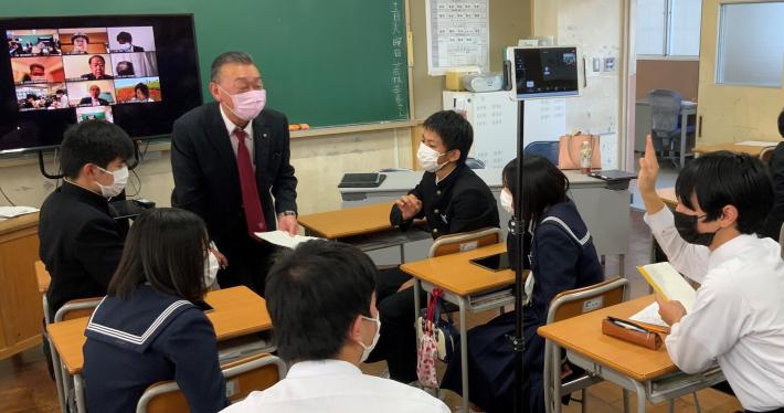 中学校の教室で、男性の議員が座っている生徒に話しかけている