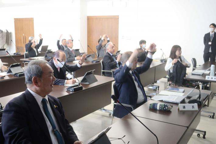 会議室で机に男性と女性の議員が座っており、一部の議員が手を挙げてジェスチャーをしている