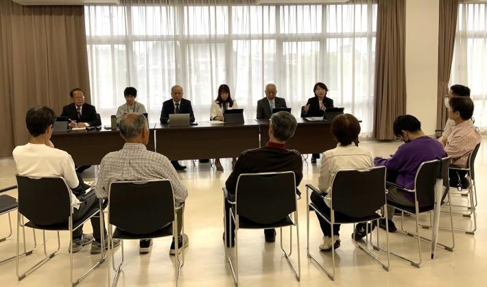 会議室で机に男性と女性の議員が座っており、その周りの椅子に参加者が座っている