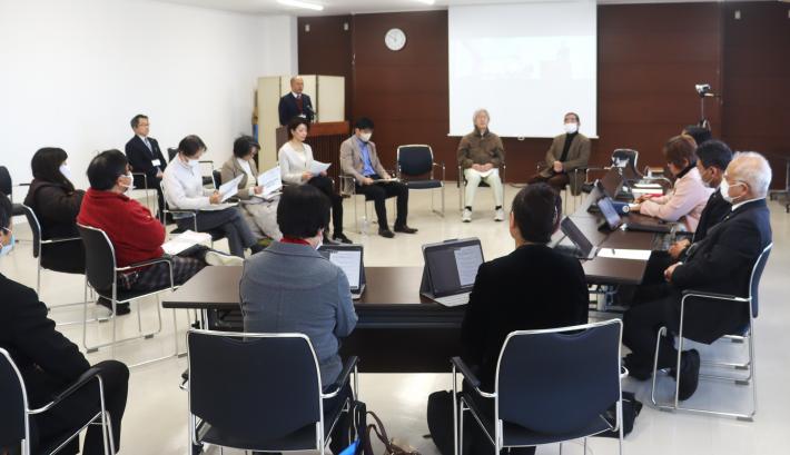 会議室で議員と市民が椅子に座って、演壇で議員が話しているのを聞いている