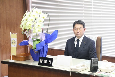 議長室内の副議長席に座る男性議員(落合副議長)。左脇には胡蝶蘭が飾られている。