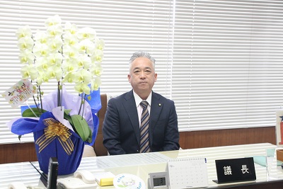 議長室内の議長席に座る男性議員（金澤議長）。机の上には胡蝶蘭が飾られている。