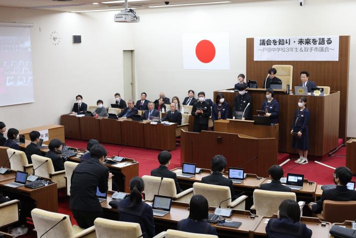 2組生徒が壇上に上がり発表している様子とその奥で発表を聞く取手市議会議員