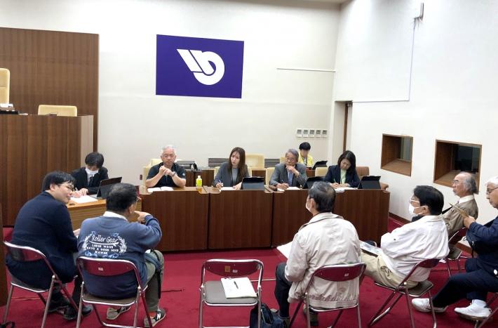 議場で議員が机に座ってメモをしながら、椅子に座っている男性数名の話を聞いている