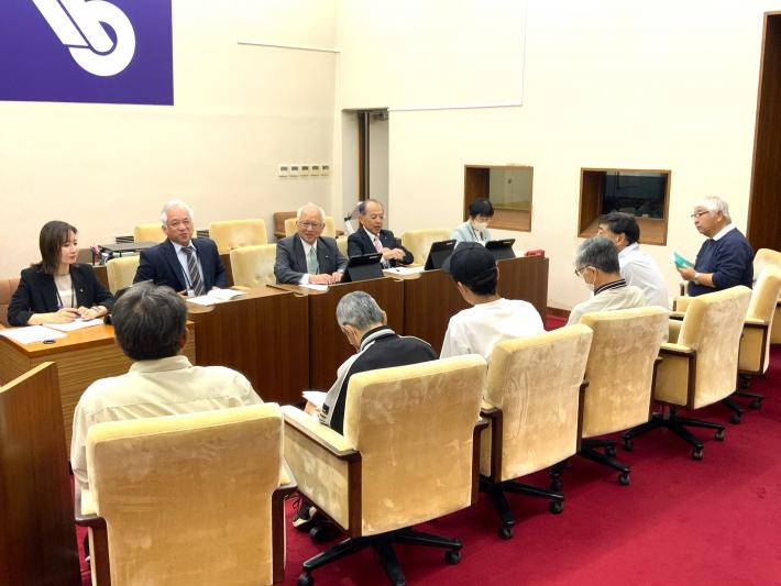 議場で男女の議員が机に座って、数名の市民から意見を聞いている