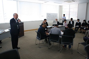 ガラス張りの会議室にて、黒いスーツの男性がマイクを持ち、笑顔で立っている。そばには男性7人と女性2人が座っている