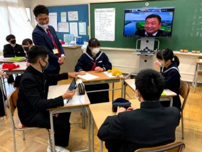 4人の生徒がグループになって着席。そのうち、男子生徒1人がタブレットを持つ。黒板前のモニターには須田議員が映る。