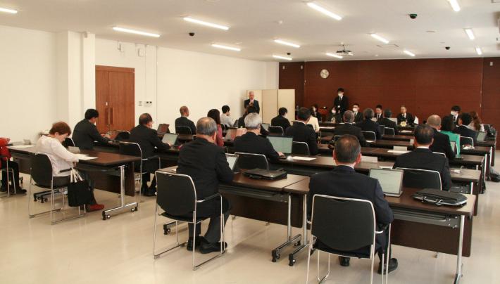 会議室内に、縦長の机が置かれ、スーツを着た男女24名がこちらに背を向けて座り、その奥にスーツを着た男女8名がいる