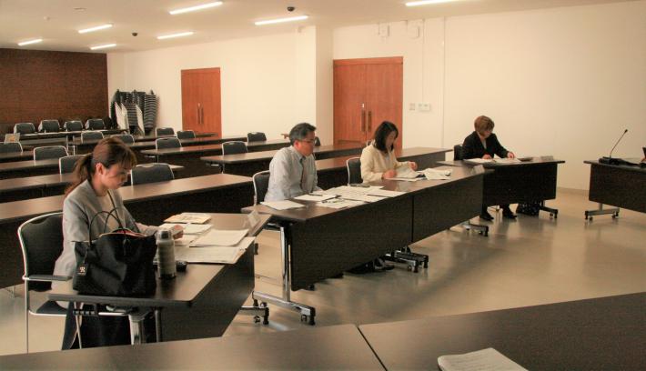 会議室内に縦長の机が4つ置かれ、スーツを着た男性と女性4名がそれぞれ座り、手元の資料を見ている