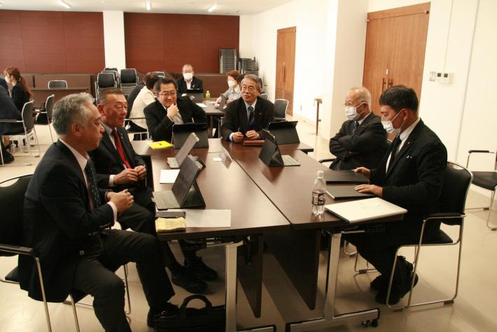 机を囲んで黒いスーツを着た短髪の男性6人が座り、議論を行っている。