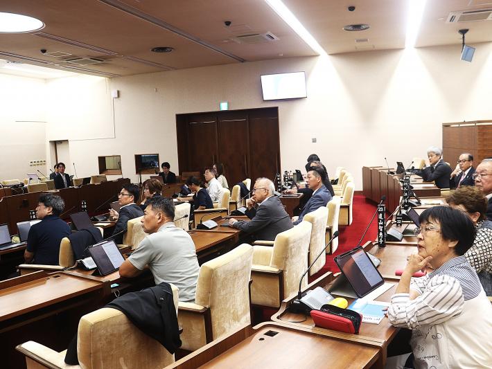 男性の研修講師が座って21名の議員に説明を行っている