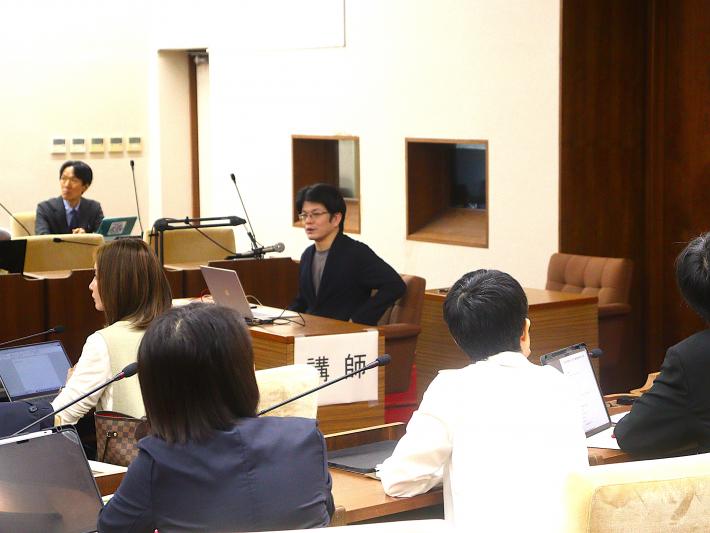 男性の研修講師が座って21名の議員に説明を行っている