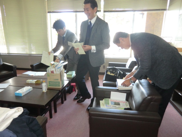 スーツを着た短髪の男性3名が会派室内に置いてある椅子やテーブルの上の書類を整理している