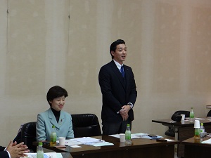 男性副委員長(関川副委員長)が起立して挨拶している。左隣に女性委員長(阿部委員長)が着席している。