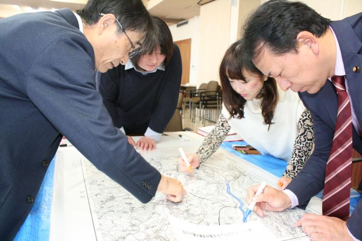 男性議員2人(結城・佐藤議員)、女性議員2人(石井・小池議員)が机の中央に置かれた地図に書き込んでいる。