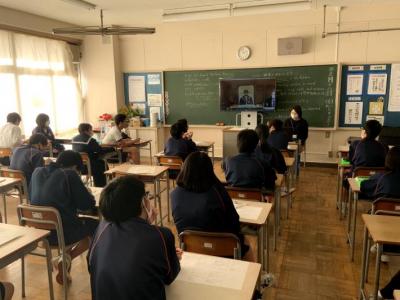 教室の黒板前に置かれたテレビを着席して見る生徒。