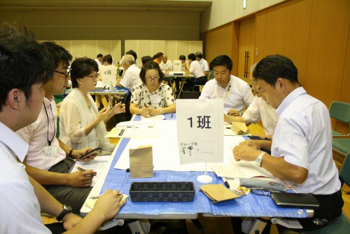 青い机を囲んで白いシャツを着た短髪の男性5名と女性2名が座っている。机上には1班とかかれた立て札が置かれている