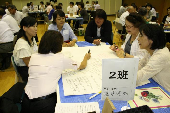 青い机を囲んでスーツを着た6名の女性が座っている。机上には模造紙と、2班と書かれた立て札が置かれている