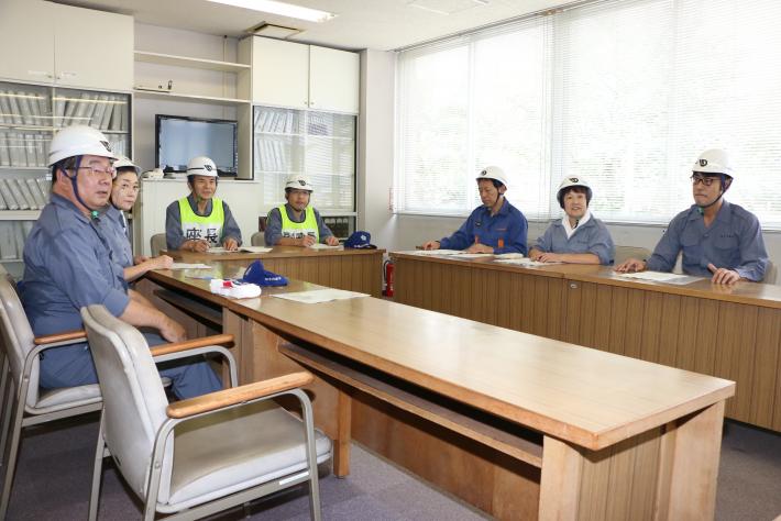 縦長の机がコの字型に置かれ、作業服を着てヘルメットをかぶった男性5名と女性2名が座っている