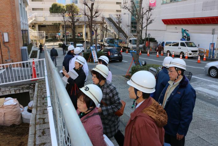 白いヘルメットを被った男性7人と女性4人がコンクリートでできた道に立って、工事の様子を見ている