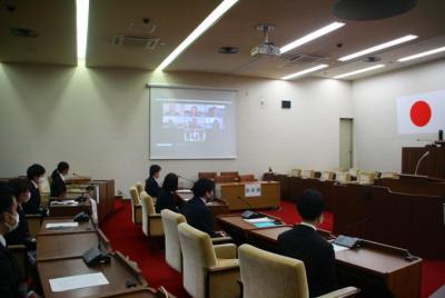 議場のスクリーンにZoomの映像が映る、8人の新規採用職員が議席に間隔を開けて座り、スクリーンを見る