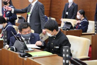 男子生徒が議席に座っている。その脇にしゃがむ男性議員(須田議員)の写真