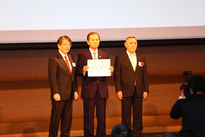 スクリーンを背景に、黒いスーツを着た男性3名が横一列に並んで立っている。中央の男性が両手で賞状を持っている