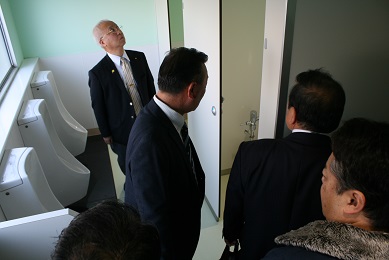 スーツを着た男性4人とジャンパーを着た男性1人が男子トイレ内に立っている