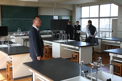 学校の理科室にスーツを着た短髪の男性4人が立っている。理科室には縦長の机が6つ置かれている