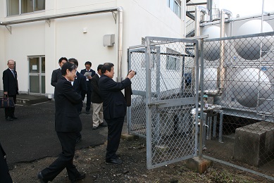 黒いスーツを着た短髪の男性7人がフェンスで囲まれた受水槽の前に立っている。手前に立っている男性がカメラで受水槽の写真を撮っている