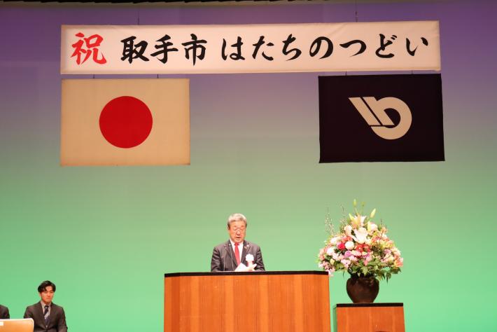 ステージ上演台にて挨拶をする市長。市長の横には壺生の花。背景には「祝 取手市はたちのつどい」の横看板の下に日本国旗と取手市旗。
