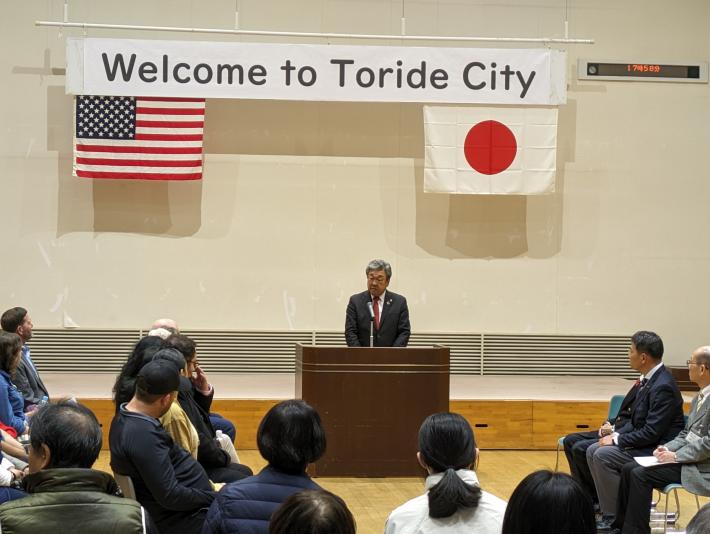 演台にて挨拶をする市長。背景には「Welcome to Toride City」の横断幕と星条旗、日本国旗