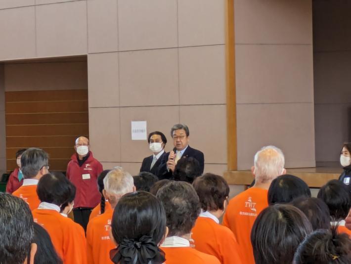 体育館ステージ前にて参加者へ挨拶をする市長。参加者はオレンジのユニフォーム姿