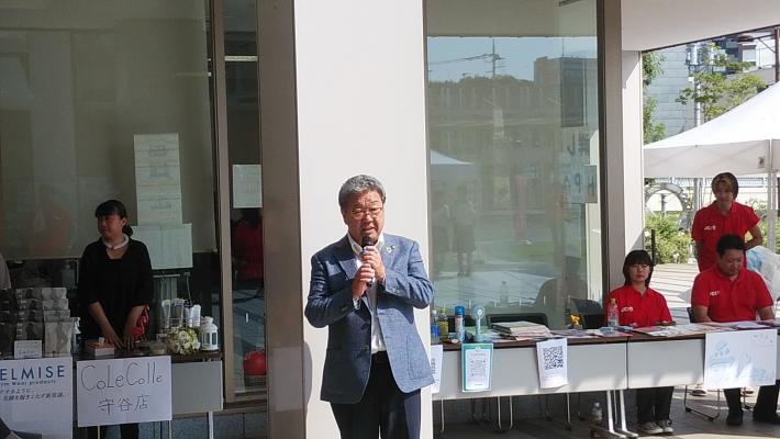 茨城南青年会議所8月例会での市長あいさつ。机が並べられ、着座の人をバックに挨拶するスーツ姿の男性。