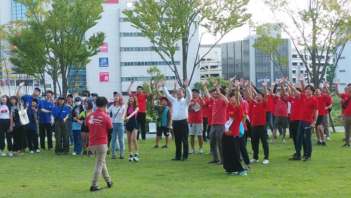 茨城南青年会議所8月例会ここがとりでだZでの集合の様子。芝生の上に赤・青のシャツを着た集団が手をもろてを上げて立っている。