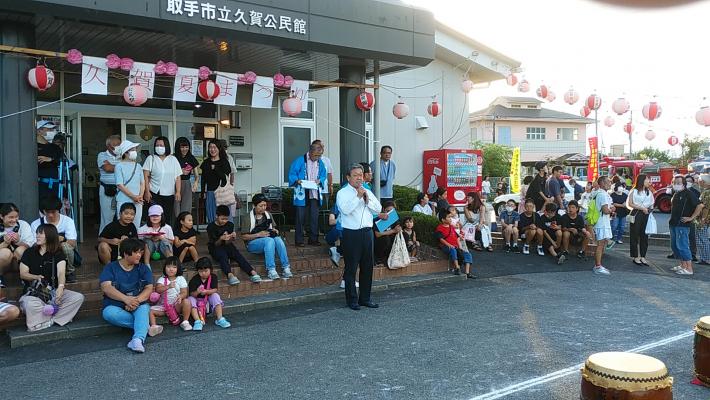 久賀公民館夏まつりでの市長あいさつ。久賀公民館の建物の前に人が大勢いて、その中央で男性が挨拶している。