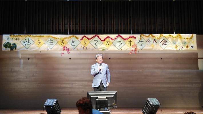 ステージ上でマイクを持って挨拶をする市長。後ろに「生き活きとりでカラオケ発表会」横断幕