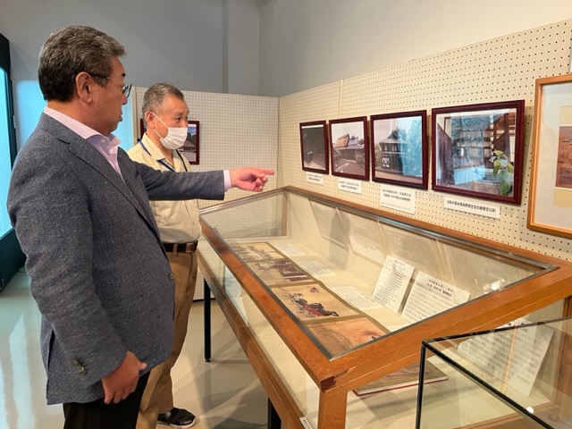 埋蔵文化財センターの展示室にて展示品を鑑賞する市長と説明をする職員。市長は壁に展示された写真を指で示している