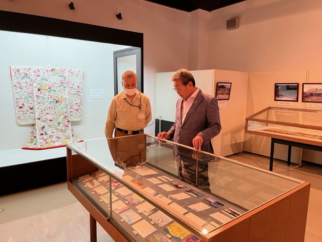 埋蔵文化財センターの展示室にて硝子ケースに展示された絵はがきを見る市長と説明をする職員