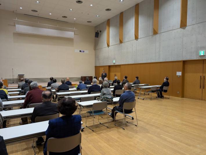 長机の前に座る人たちの前であいさつをする男性