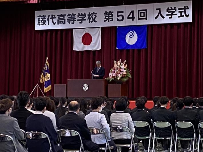 ステージで祝辞を述べる男性とそれを聞く人たち