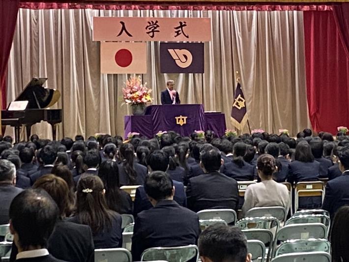 ステージで祝辞を述べる男性とそれを聞く人たち