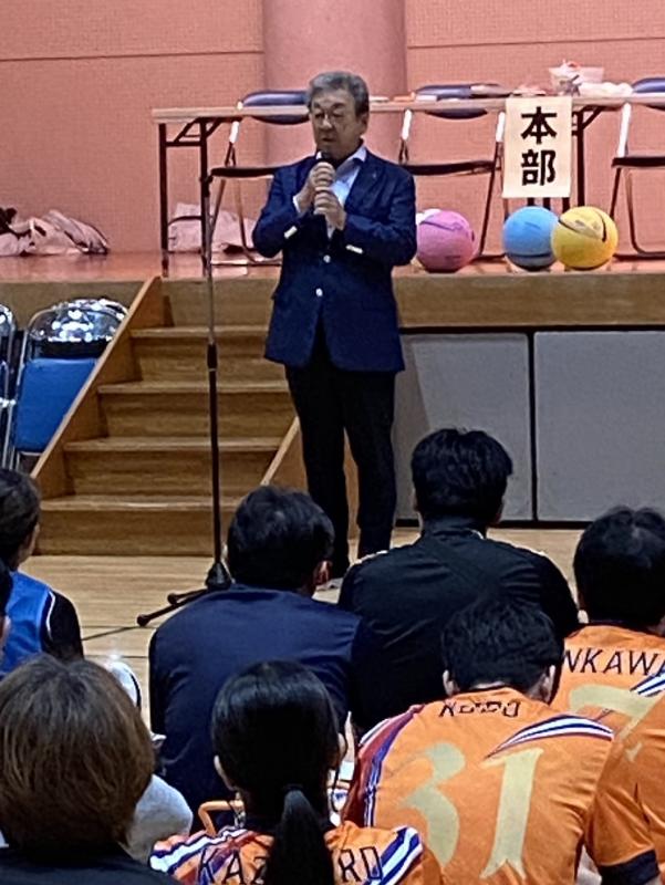体育館の床に座る参加者に向けて話す、スーツを着た男性