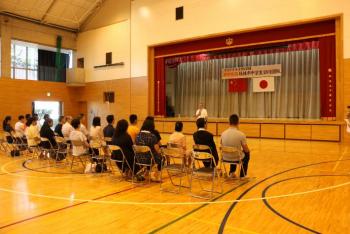 取手二中体育館でのウェルカムセレモニー。舞台上に両国国旗と歓迎横断幕が掲げられ、男性が着席した両国関係者に挨拶をしている。