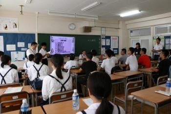 黒板前のホワイトボードを使って男子生徒が発表を行い、それを10人以上の日中両国中学生が着席で聞く様子を教室後方から撮影した写真。